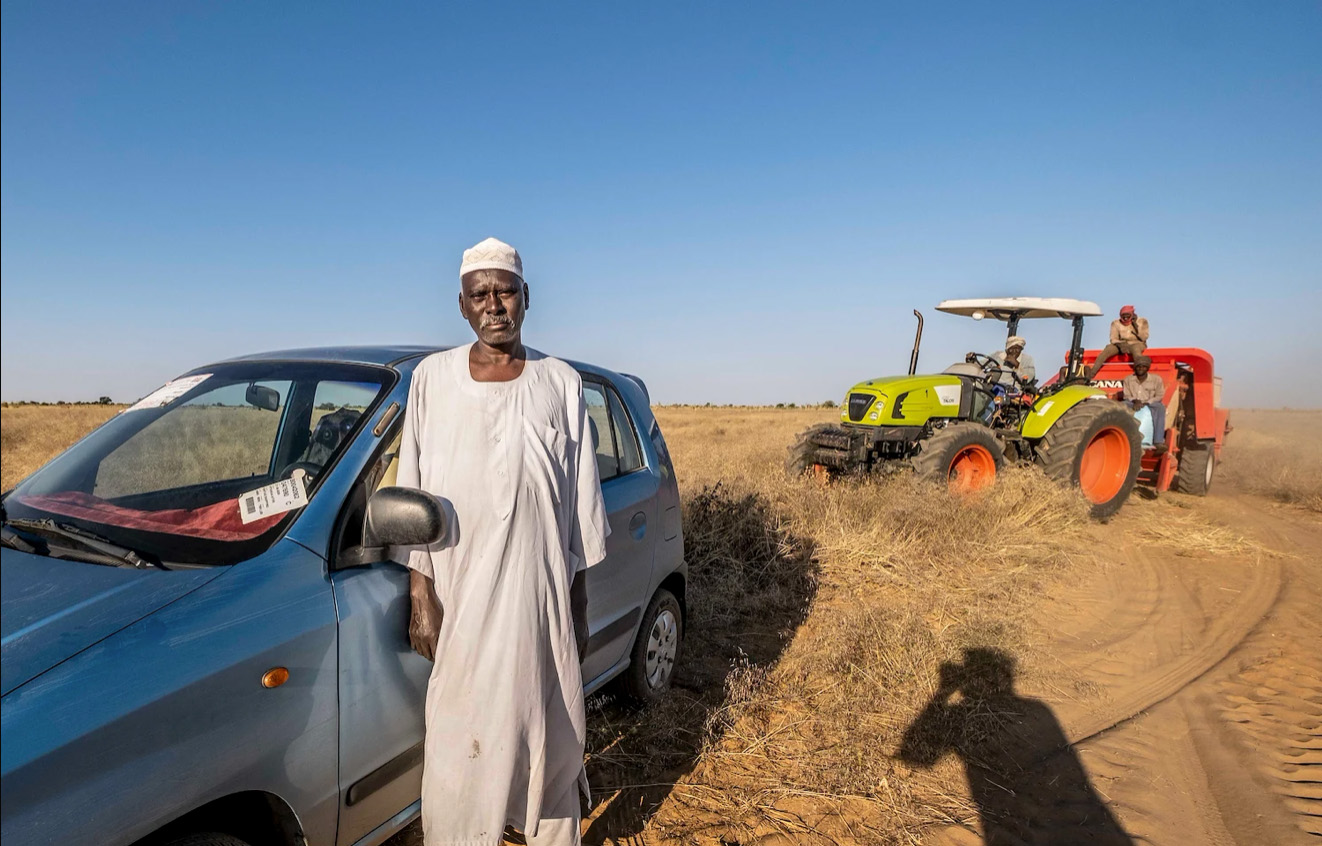 Photo of a Sudanese man standing next to a car with a tractor in the background